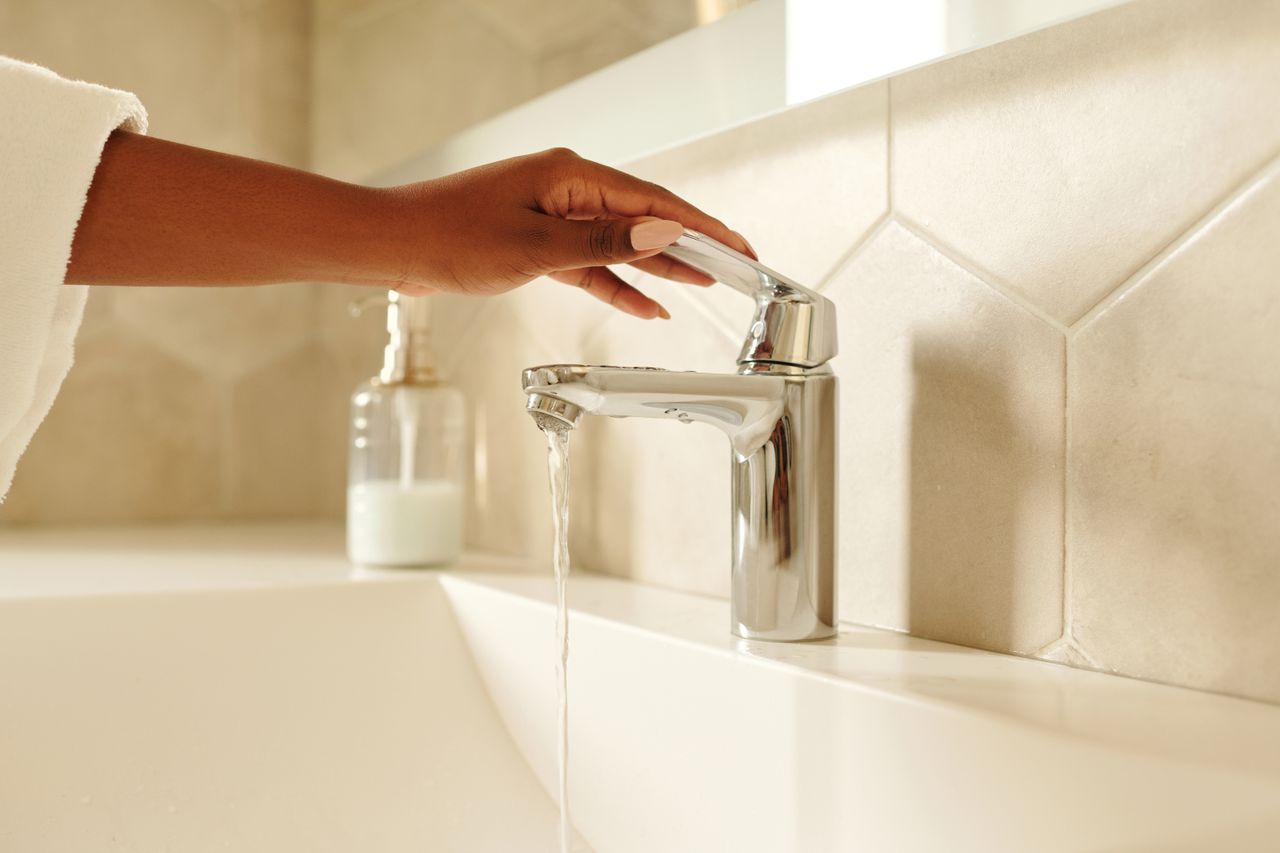about-02 A close-up shot of a hand operating a chrome bathroom faucet with water flowing in a modern bathroom setting.